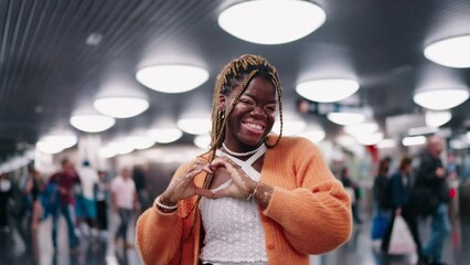 young African American woman with braided hair and vitiligo making heart shape with hands and smiling warmly. background blurred with people walking by, concept of love and connection.
