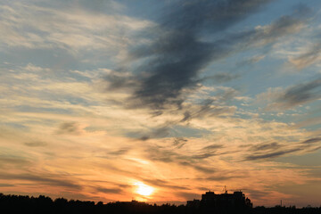 Blue evening sky with rare white clouds, evening twilight, sun through the clouds over the black silhouette of an industrial city