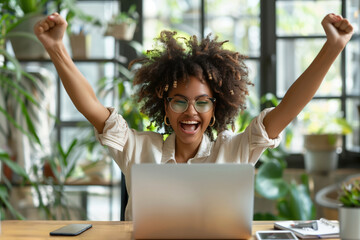 An ecstatic young woman with curly hair and glasses raises her arms in a victory pose in front of her laptop, surrounded by houseplants. Goal accomplished, project finished. - obrazy, fototapety, plakaty