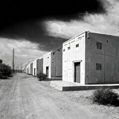 Fototapeta na wymiar Outdoor black and white photo of a row of industrial mini warehouses along a gravel road, no traffic, cloudy sky, light and shadow, empty. From the series “Terminal Beach.