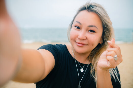 Young asian pretty woman shows heart sign making selfie with happy smile with ocean beach on background
