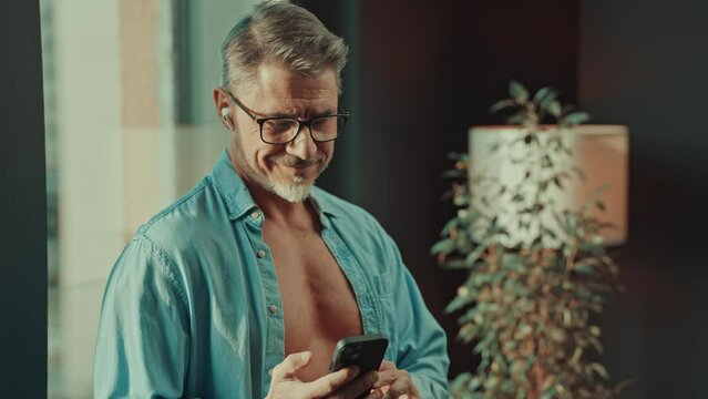 Mid adult man standing in sunlight in open shirt in the morning, checking email, news or social media on cell phone. Happy older male smiling in glasses.