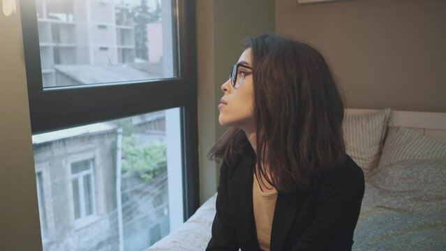 Young woman gazing out bedroom window, contemplative mood, city backdrop.