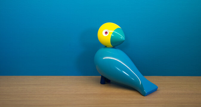 A colorful toy bird made of wood sitting on a shelf in front of a blue wall. The head of the bird is yellow. 
