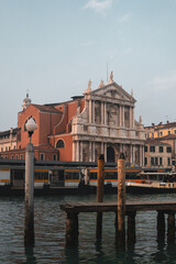 View from Fondamenta San Simeon Piccolo: Old wooden pier on Grand Canal, water bus station, Santa Maria di Nazareth church, surrounded by colorful Venetian buildings. Blue sky over the iconic scene.