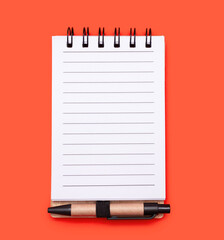 Open spiral notebook in a line, with a small pen on a orange background. A small notebook with blank white lined pages and a pen. Free space for text and list