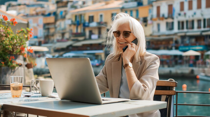 Blonde elderly woman in a cafe on the waterfront near the marina, working online, talking on a smartphone with business partners during lunch - obrazy, fototapety, plakaty