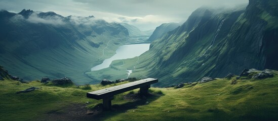 A bench is perched atop a grassy highland, offering a scenic view of a mountain valley with clouds hovering above. The natural landscape is accentuated by the atmospheric phenomenon in the sky - obrazy, fototapety, plakaty