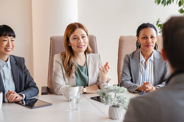 A diverse group of multi-ethnic businesswomen sit in a corporate office and have a meeting with a business associate - 761818883