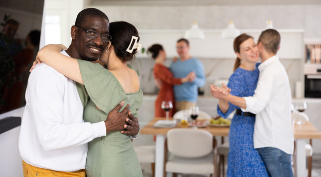 Smiling black man in casual white sweater tenderly embracing woman in green off-shoulder dress, enjoying slow dance at cozy friendly house party