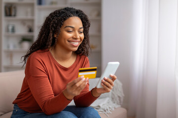 Smiling black woman at home holding credit card and smartphone, shopping online - obrazy, fototapety, plakaty