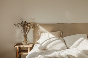 an unmade bed with white linen and a beige headboard, soft sunlight streaming through the window casting gentle shadows on the wall behind it. A vase holding dried flowers sits beside the bed.