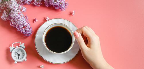 Female hands holding cup of espresso coffee. Alarm clock and lilac flowers on light background.