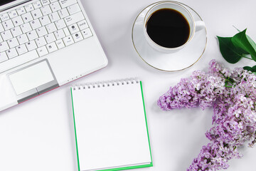 Spring flatlay. Notepad, laptop, lilac flowers and cup of coffee on light background.