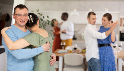 Fototapeta premium Smiling middle-aged man in blue jumper and glasses enjoying slow dance with brunette in olive dress at relaxed friendly home gathering