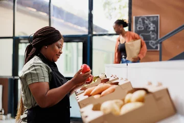 Poster Female storekeeper checking and arranging freshly harvested produce on shelves. Black woman wearing an apron holding and examining red apples in boxes at local convenience store. © DC Studio