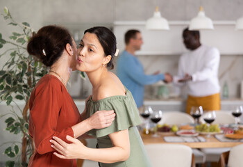 Obraz na płótnie Canvas Smiling Asian woman welcoming arriving friends for informal dinner with food and drinks served on table in background, giving greeting kiss on cheek to bestie..