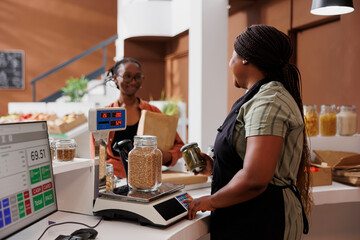 An aproned black woman assists a customer at eco friendly store counter with weighing local,...