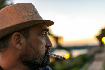 Handsome Mature Man with Beard and Hat Smoking a Cigarette During a Sunset in the City