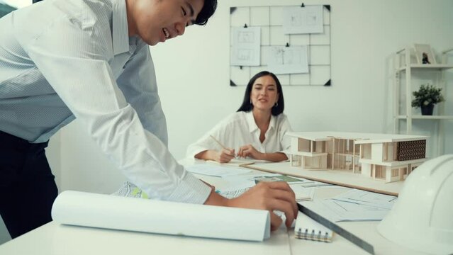 Skilled caucasian interior designer working with house model while discussing with asian architect engineer about house plan on meeting table with blueprint and house model scatter around. Immaculate.