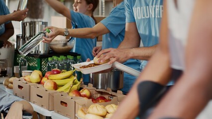At food drive, poor female wheelchair user receives free food and provisions. Multiethnic volunteers in blue t-shirt distributing fresh fruits and hot meals to less privileged. Close-up, tripod. - obrazy, fototapety, plakaty
