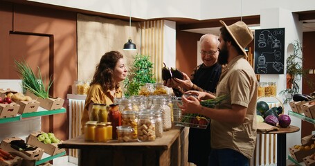 Senior vendor assisting clients in eco friendly shop with ethical sourced organic food items, providing recommendations. Couple in local store chatting with trader about pantry staples bulk products - obrazy, fototapety, plakaty