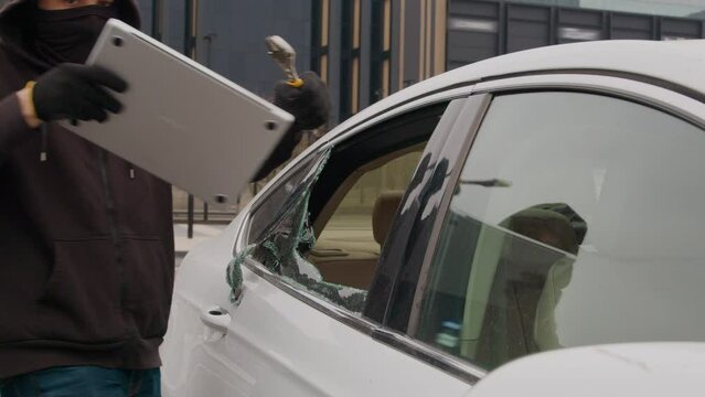 A thief in a black hoodie steals a laptop through the broken window of a car parked in a shopping mall, showing a daring act of carjacking