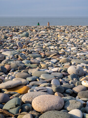Gray sand and stones on the beach. Two silhouettes of people on the shore in the distance. Beach in winter. Sea coast soil.