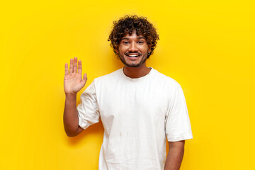 young cheerful indian man in white t-shirt waving and greeting on yellow isolated background, curly guy showing palm gesture welcome and goodbye © Богдан Маліцький