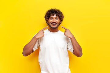 young cheerful indian man in white t-shirt pointing white toothy smile on yellow isolated...