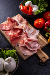 cutting board with mixed cold cuts, vegetables and mozzarella on a black background