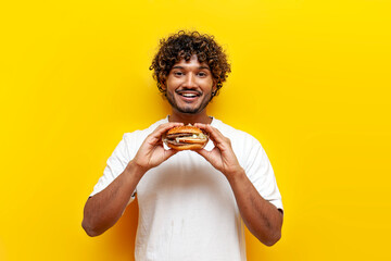 young indian man holding tasty burger and smiling on yellow isolated background, curly guy student...