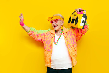 funny crazy granny in hipster clothes listening to music on a tape recorder and showing a kiss on a yellow isolated background, elderly cool woman dancing at a party and having fun - obrazy, fototapety, plakaty