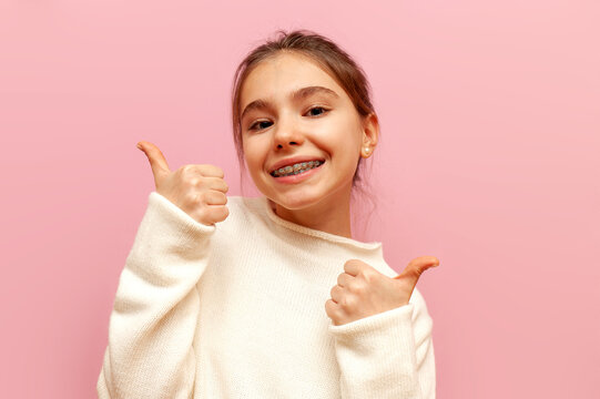 cheerful teenage girl with braces smiles and shows likes with her hands on a pink isolated background, child shows thumbs up and recommends