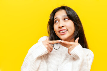 young shy asian woman with braces feeling shy and daydreaming over yellow isolated background,...