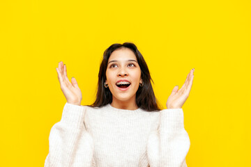 young shocked asian woman with braces wonders and holds empty hands over yellow isolated background, korean girl raises her hands up in amazement - obrazy, fototapety, plakaty