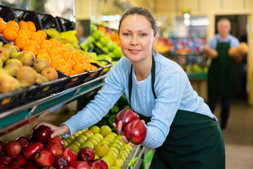 Diligent middle-aged shop assistant laying fresh apples on counter in big greengrocery