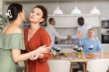 Women greet each other with kiss on cheek before festive dinner. Men shaking hands in the background