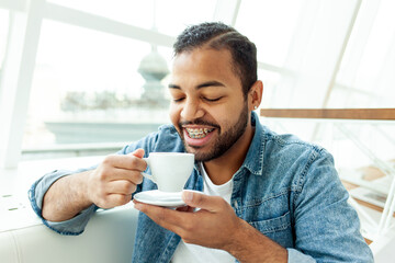 African American man with braces drinks coffee from a cup and smiles in a white cafe, a man with stubble drinks tea from a white mug - obrazy, fototapety, plakaty