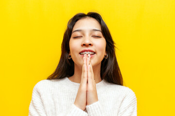 young cheerful asian girl with braces praying and asking for help on yellow isolated background,...