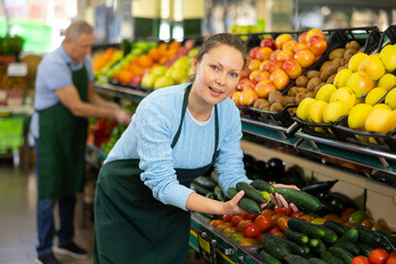 female seller of vegetable shop chooses fresh and ripe cucumbers for regular customer