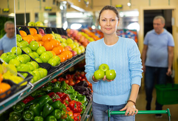 Happy middle-aged female in casual clothes shopping and choosing green apples in supermarket
