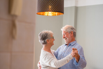 Joyful active old retired romantic couple dancing laughing in living room, happy middle aged wife and elder husband having fun at home, smiling senior family grandparents relaxing bonding together.