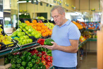 elderly man at vegetable showcase of supermarket ponders - to buy green or red bell peppers