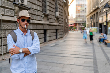 Handsome Well Dressed Young Man with Hat, Sunglasses and Backpack Standing in the City Center and...