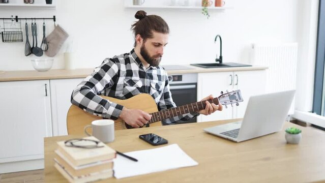 Portrait of handsome bearded adult posing with six-string guitar while enjoying pleasant pastime at home. Smiling Caucasian man learning to play instrument using online lessons on laptop.