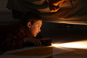 Little girl with flashlight looking for monster under bed at night