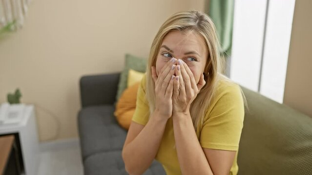 Cheeky blonde lass in a t-shirt, suppressing a laugh at home, hand over her mouth, caught up in juicy gossip and scandal! trembles with fun, embarrassment on her radiant face.