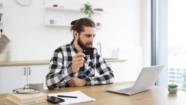 Joyful Caucasian man in casual clothes using credit card while running portable computer in workplace. Smiling bearded person in checkered shirt enjoying virtual shopping using online wallet from home