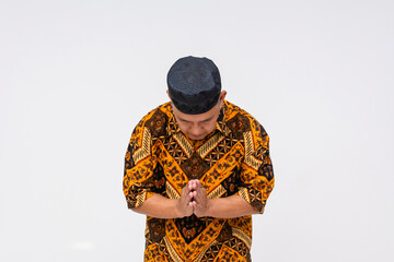 Humble and respectful Indonesian man in batik shirt and kopiah hat bowing with hands together isolated on a white background.